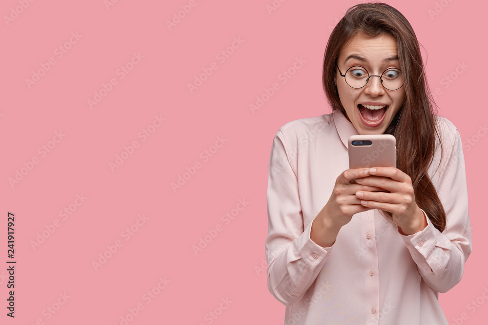 Studio shot of pretty woman stares at mobile phone, feels satisfied, stares at screen, opens mouth, dressed in formal shirt, isolated over pink background with blank space for your promotion Studio shot of pretty woman stares at mobile phone, feels satisfied, stares at screen, opens mouth, dressed in formal shirt, isolated over pink background with blank space for your promotion