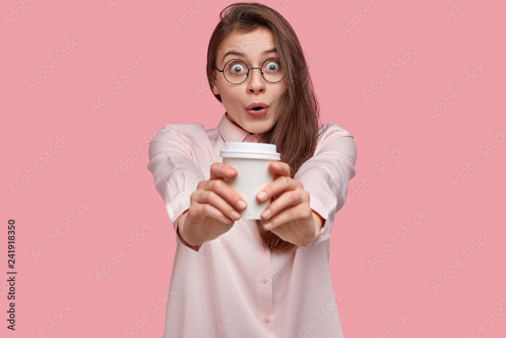 Amazed beautiful young woman with surprised gaze stretches hands with takeaway coffee, dressed in formal shirt, keeps jaw dropped, dressed elegantly, enjoys drinking hot beverage, has spare time Amazed beautiful young woman with surprised gaze stretches hands with takeaway coffee, dressed in formal shirt, keeps jaw dropped, dressed elegantly, enjoys drinking hot beverage, has spare time