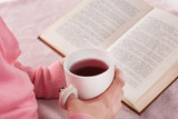 Woman holding cup of hot tea drink and reading book at home in bed. Girl wears pink woolen sweater. Education and cold weather concept. Close up, selective focus