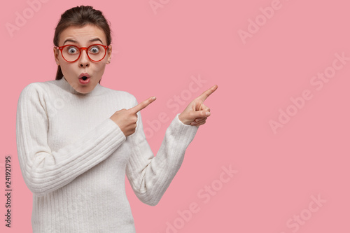 Studio shot of surprised emotive woman feels astonished, points with both index fingers, has bated breath, dressed in white sweater, models against pink background, involved in advertising campaign