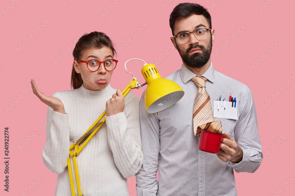 Unaware hesitant young woman and man have clueless expressions, stand next to each other, carry yellow desk lamp, cup of drink, wear spectacles for vision correction, isolated over pink background Unaware hesitant young woman and man have clueless expressions, stand next to each other, carry yellow desk lamp, cup of drink, wear spectacles for vision correction, isolated over pink background
