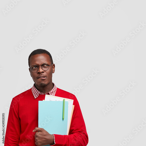 Black confused male worker with thoughtful nervous facial expression, bites lower lip, carries textbooks, wears spectacles and red sweater, stands over white studio wall with blank space for your text