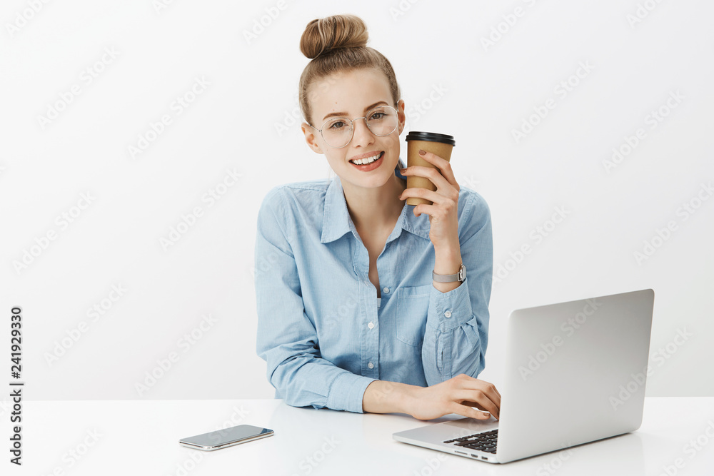Charming young female manager in office sitting near laptop raising cup of coffee and smiling at camera working late talking to coworker during break discussing recent rumors over gray wall Charming young female manager in office sitting near laptop raising cup of coffee and smiling at camera working late talking to coworker during break discussing recent rumors over gray wall