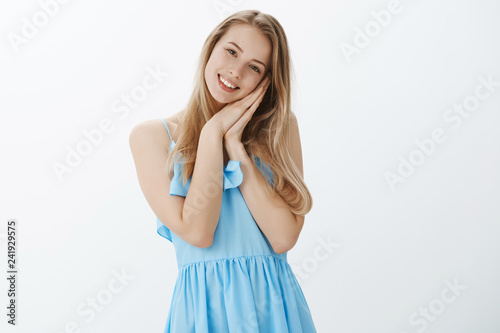 Dreamy sweet girlfriend feeling happy leaning head on palms pressed together as if pillow smiling broadly being touched with cute gift standing in blue elegant dress over gray background