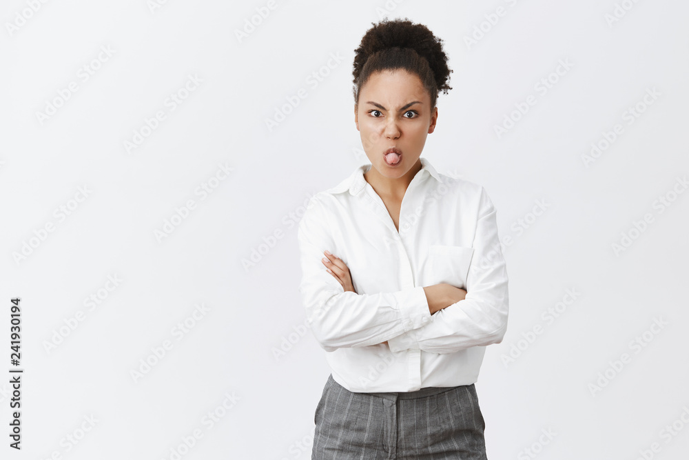 Girl being moody, showing her charachter while having silly argument with girlfriend, holding hands crossed on chest, sticking out tongue and frowning, misbehave and bieng childish over grey wall Girl being moody, showing her charachter while having silly argument with girlfriend, holding hands crossed on chest, sticking out tongue and frowning, misbehave and bieng childish over grey wall