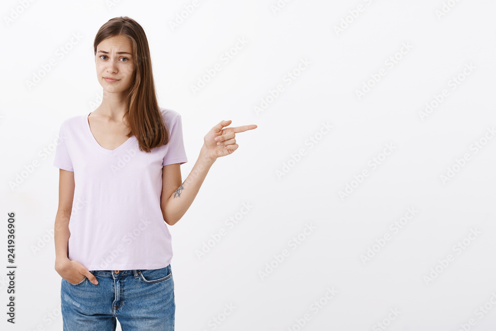 Woman feeling doubtful and uncertain about place friend wants visit. Unsure displeased attractive and smart female brunette pointing right pursing lip and frowning from dislike over white wall Woman feeling doubtful and uncertain about place friend wants visit. Unsure displeased attractive and smart female brunette pointing right pursing lip and frowning from dislike over white wall
