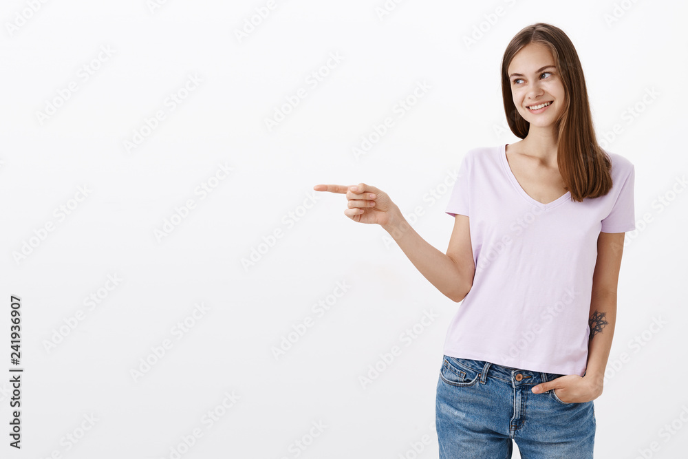 Studio shot of delighted cute caucasian female brunette with awesome tattoo on arm pointing and looking left with appreciation and thankful expression smiling joyfully posing against white wall Studio shot of delighted cute caucasian female brunette with awesome tattoo on arm pointing and looking left with appreciation and thankful expression smiling joyfully posing against white wall