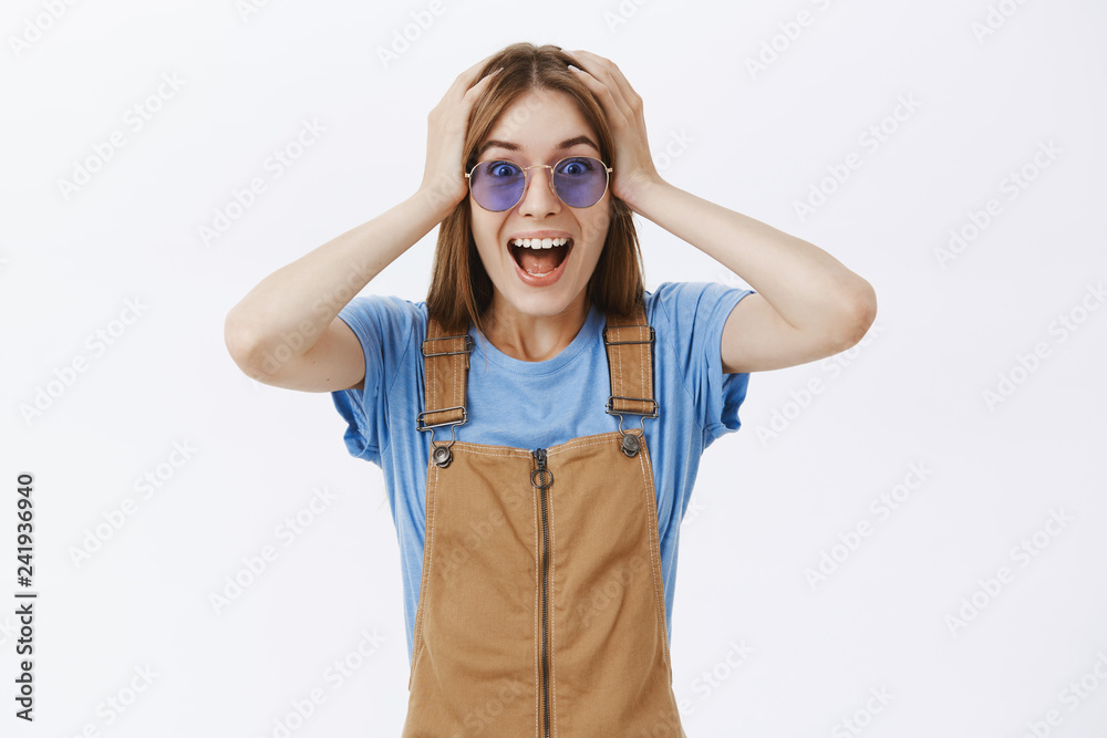 Head going round from happiness and amazement. Portrait of joyful delighted cheerful young woman in brown overalls and sunglasses holding hands on hair yelling from joy and excitement over grey wall Head going round from happiness and amazement. Portrait of joyful delighted cheerful young woman in brown overalls and sunglasses holding hands on hair yelling from joy and excitement over grey wall