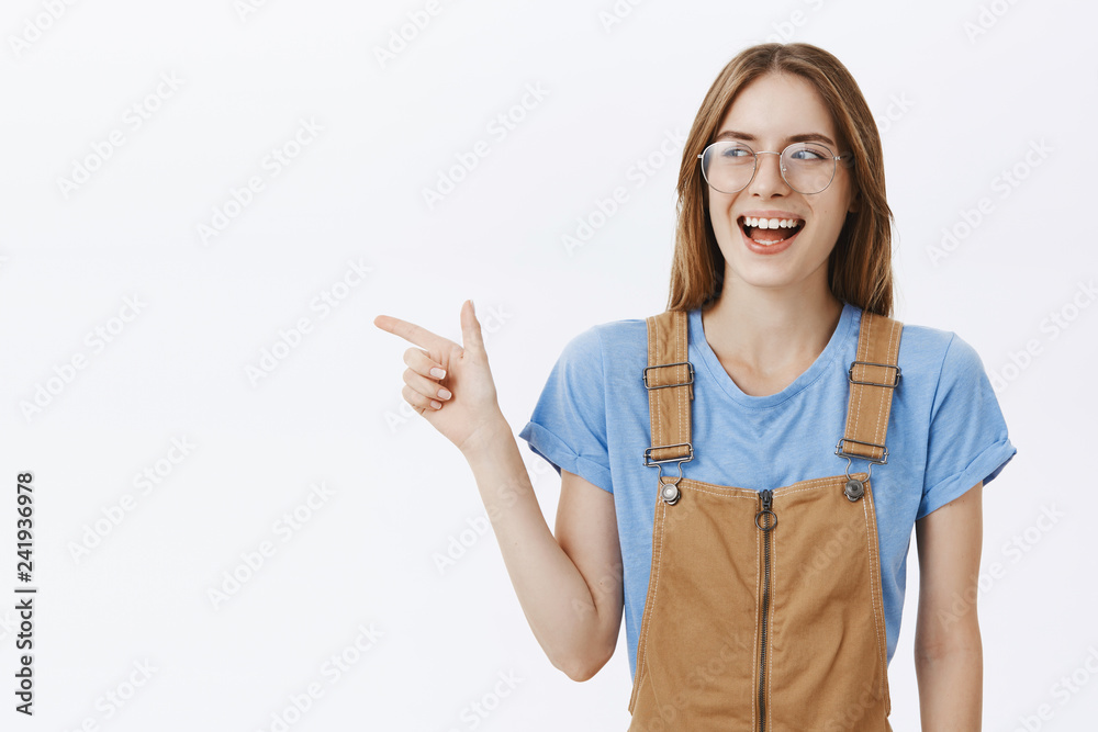 Waist-up shot of enthusiastic charming and charismatic happy girlfriend in brown overalls and transparent glasses pointing and looking left joyfully laughing having fun against gray background Waist-up shot of enthusiastic charming and charismatic happy girlfriend in brown overalls and transparent glasses pointing and looking left joyfully laughing having fun against gray background