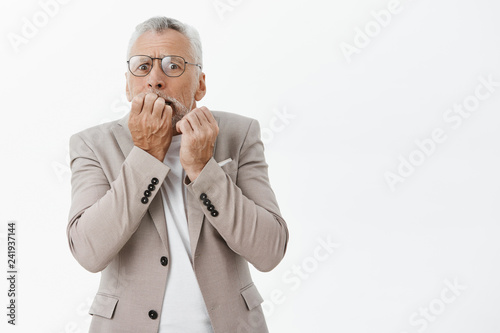 Old rich man scared losing money. Portrait of nervous afraid senior male model in glasses and suit biting fingers from anxiety and fear panicking staring terrified at camera against white background © Cookie Studio