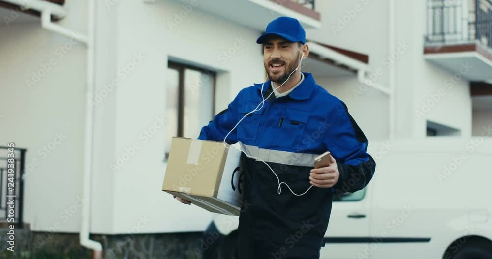 Cheerful and attractive Caucasian deliveryman in the blue uniform walking with a parcel in hands and dancing as listening to the music un headphones. Outdoor.