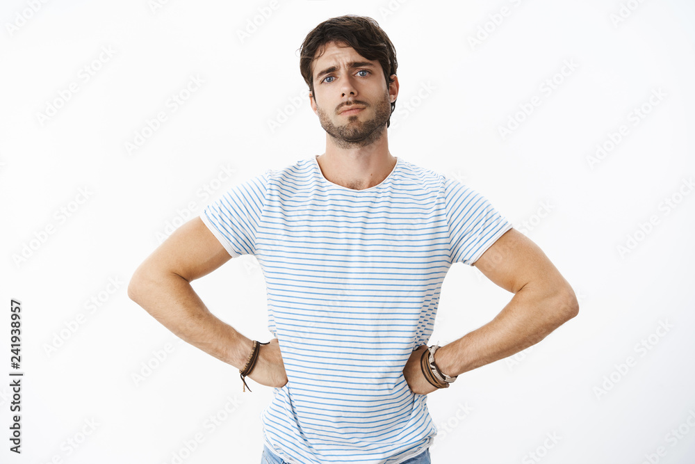 Unretouched shot of handsome european man with beard and blue eyes feeling upset and disappointed as friend broke promise frowning and sulking holding hands on waist in scolding gesture, judging Unretouched shot of handsome european man with beard and blue eyes feeling upset and disappointed as friend broke promise frowning and sulking holding hands on waist in scolding gesture, judging