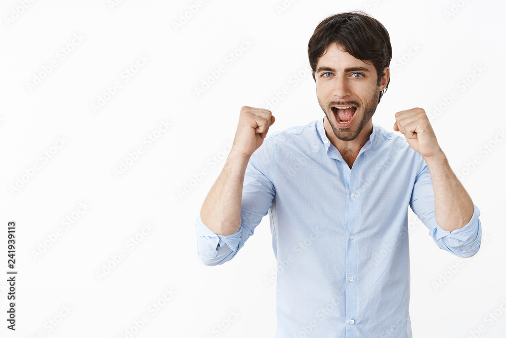 Joyful excited good-looking entrepreneur cheering and triumphing yelling yes and raising clenched fists in victory and sucess gesture rejoicing from good news standing pleased over grey wall Joyful excited good-looking entrepreneur cheering and triumphing yelling yes and raising clenched fists in victory and sucess gesture rejoicing from good news standing pleased over grey wall