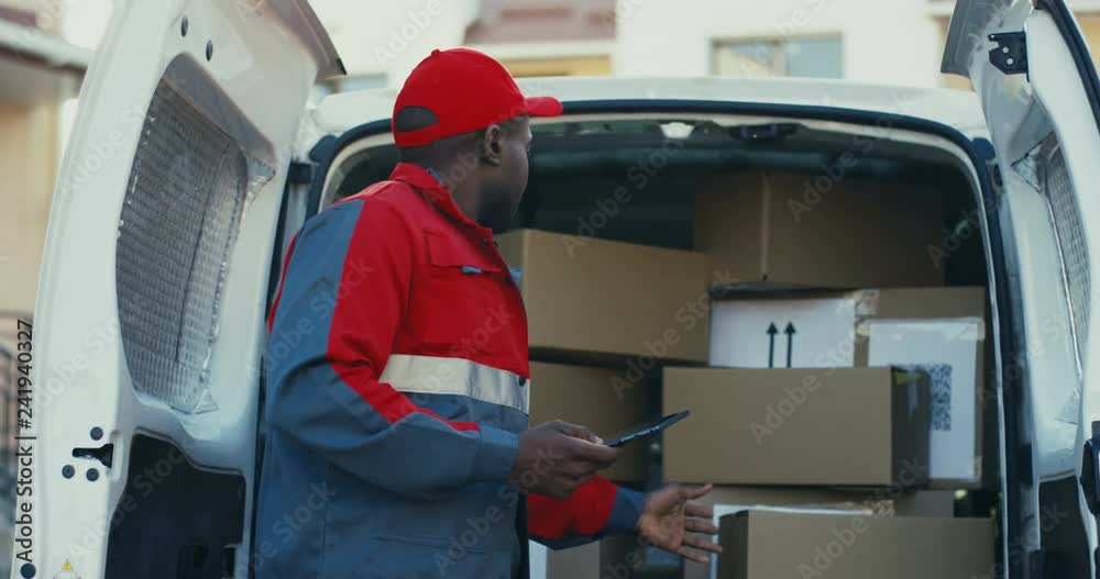 Young African American man worker of mail company counting and checking parcels in the van with tablet device in hands. Outdoor. Young African American man worker of mail company counting and checking parcels in the van with tablet device in hands. Outdoor.