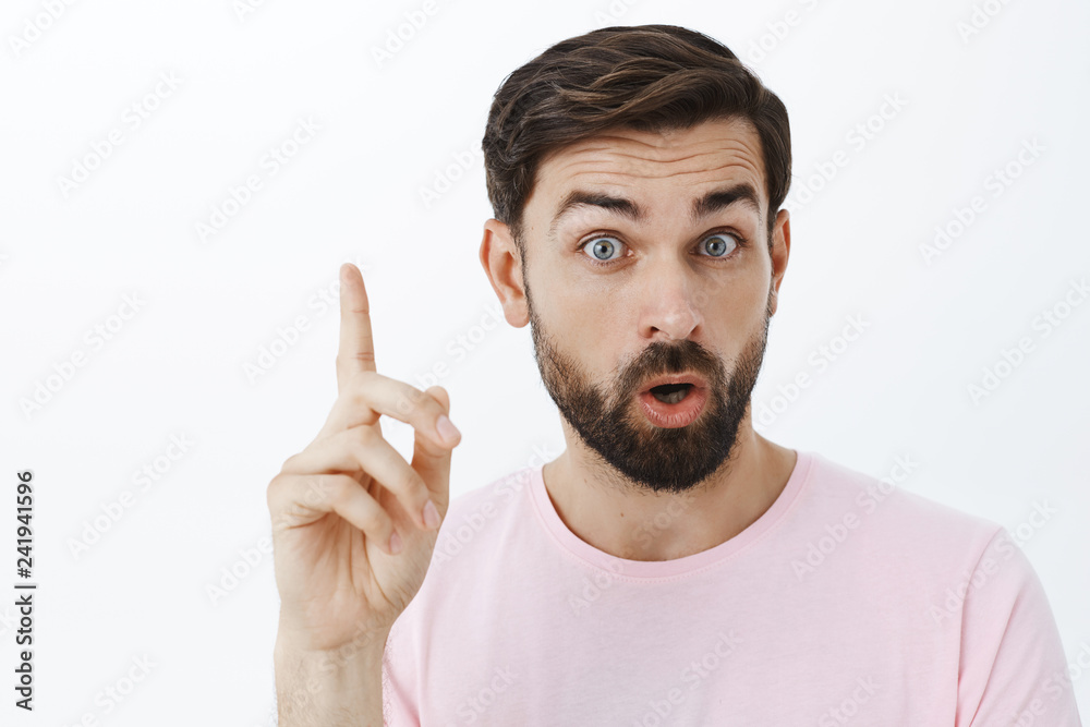 Close-up shot of creative and excited handsome male with beard and short dark hair open mouth raising eyebrow showing index finger in eureka gesture while talking adding suggestion against gray wall Close-up shot of creative and excited handsome male with beard and short dark hair open mouth raising eyebrow showing index finger in eureka gesture while talking adding suggestion against gray wall