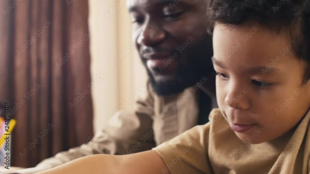 Tilt up shot of concentrating biracial little boy sitting near his father, scribbling with felt pen on paper and grimacing Tilt up shot of concentrating biracial little boy sitting near his father, scribbling with felt pen on paper and grimacing