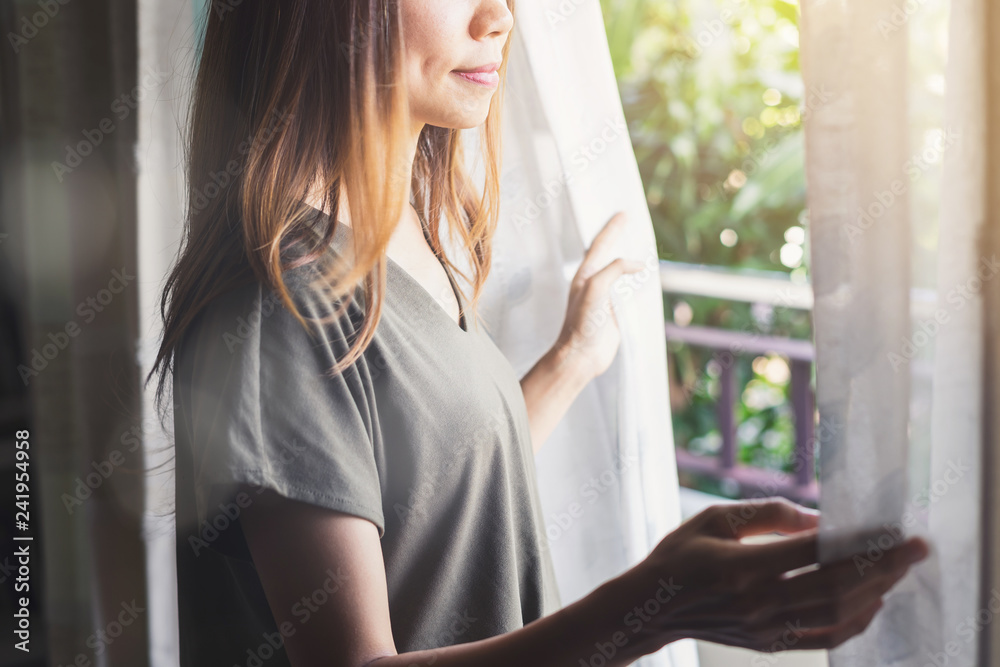 Young asian woman unveil curtain and looking out of window in the morning Young asian woman unveil curtain and looking out of window in the morning