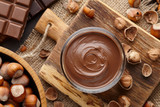 Chocolate spread or nougat cream with hazelnuts in glass jar, bowl of nuts and chocolate bars on brown countertop