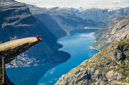 A man in a red jacket sitting on the Trolltunga rock with a blue lake 700 meters lower and interesting sky with clouds