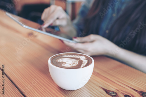 white coffee cup with latte art heart pattern on a wooden table background and Young woman hand holding smartphone in a coffee shop cafe