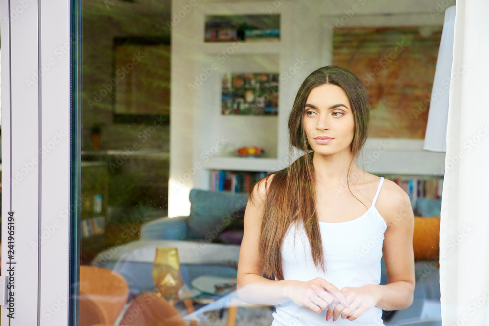 Beautiful young thinking woman looking away while standing behind the window at home Beautiful young thinking woman looking away while standing behind the window at home