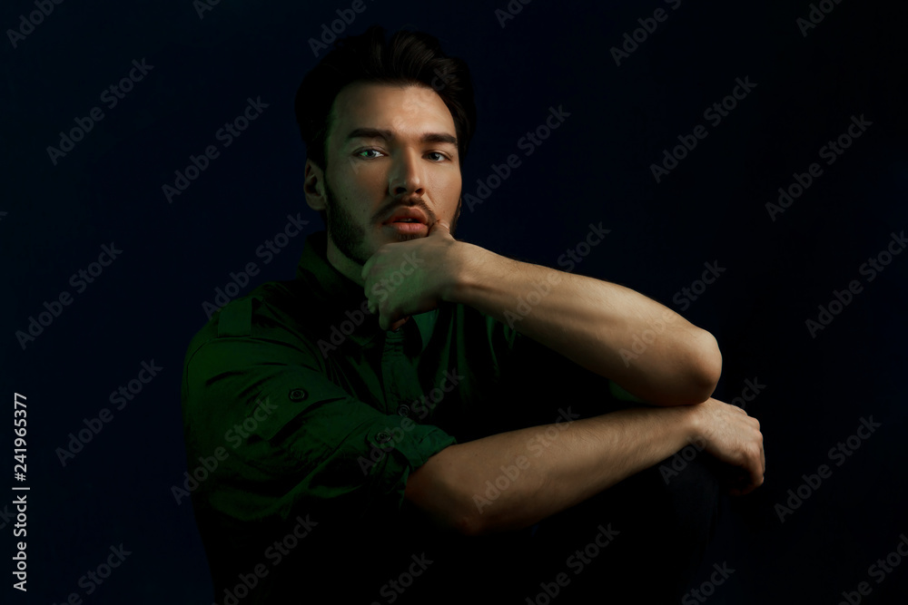 Fashion dark style portrait of young man in green shirt and style haircut poses over dark blue wall with green contrast color light