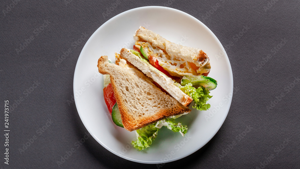 Homemade sandwich made of toast bread, fried egg, melted cheese, tomatoes, cucumber, lettuce, burgers and mayonnaise sauce. Sandwich on a white plate, on a black background. top view