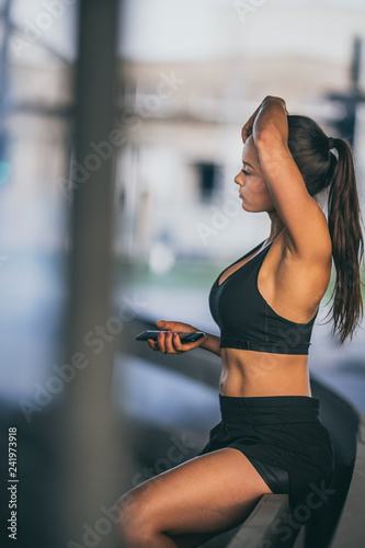 Beautiful Confident Fitness Girl in Black Athletic Top is Using a Smartphone on a Street. She is in an Urban Environment Under a Bridge with Cars in the Background.