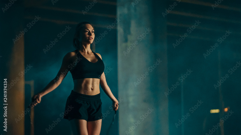 Beautiful Energetic Fitness Girl in Black Athletic Top and Shorts is Skipping/Jumping Rope. She is Doing a Workout in an Evening Foggy Urban Environment Under a Bridge.