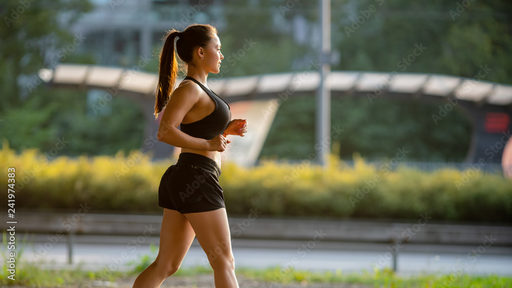 Beautiful Busty Fitness Girl in Black Athletic Top and Shorts is  Energetically Running in the Street. She is Jogging in an Urban Environment  Under a Bridge. Stock Photo