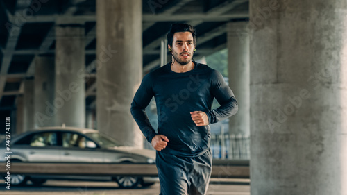 Athletic Young Man in Earphones and Sports Outfit is Jogging in the Street. He is Running in an Urban Environment Under a bridge with Cars in the Background.