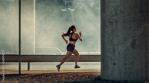Beautiful Fitness Girl in Black Athletic Top and Shorts is Energetically Running in the Street. She is Jogging in an Urban Environment Under a Bridge with Cars in the Background.