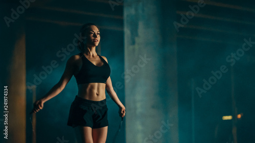Beautiful Energetic Fitness Girl in Black Athletic Top and Shorts is Skipping/Jumping Rope. She is Doing a Workout in an Evening Foggy Urban Environment Under a Bridge.