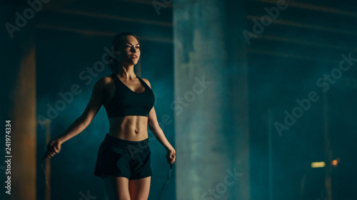 Beautiful Energetic Fitness Girl in Black Athletic Top and Shorts is Skipping/Jumping Rope. She is Doing a Workout in an Evening Foggy Urban Environment Under a Bridge. © Gorodenkoff