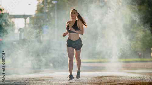 Beautiful Fitness Girl in Black Athletic Top and Shorts is Energetically Running in the Street. She is Jogging in an Urban Environment with Blurred Cars in the Background.