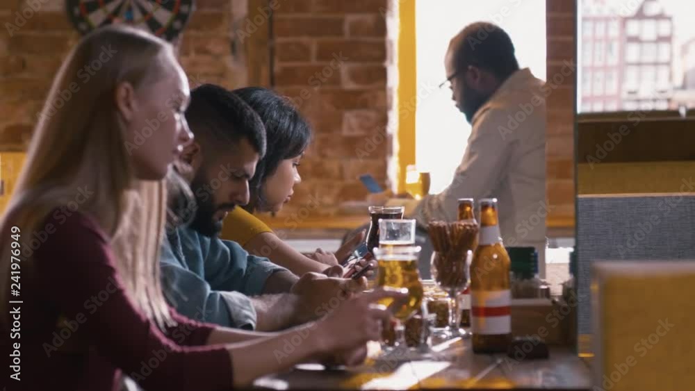 Panning shot of young people sitting at bar counter with glasses of beer and doing telework with their mobile phones Panning shot of young people sitting at bar counter with glasses of beer and doing telework with their mobile phones