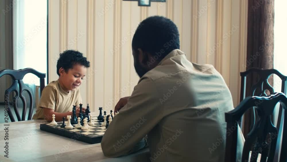 Over-the-shoulder shot of son studying to play checkmate with father at home and smiling