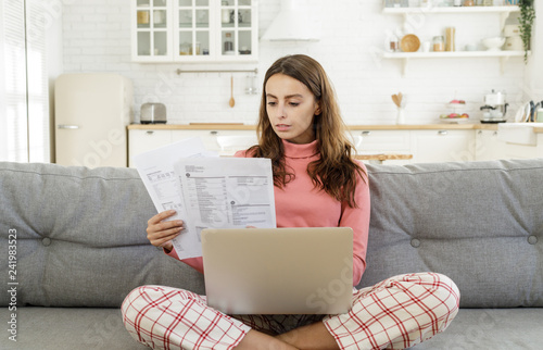 Young woman siiting on sofa in living room with laptop open and bills checking details of payment that should be made photo