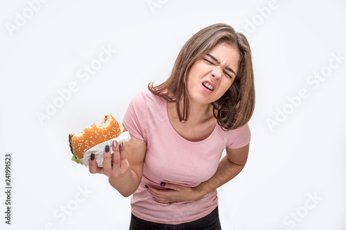 Young woman suffer from pain in stomach. She hold bitten burger. Another hand is on stomach area. Isolated on white background. photo