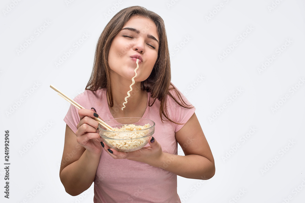 Joyful young woman eat vermicelli. She hold bow with it and fork in hands. Isolated on white background.