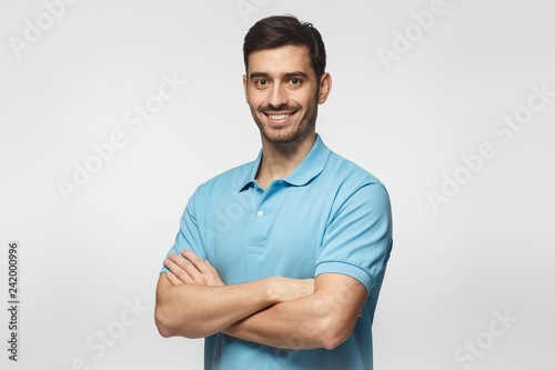 Portrait of attractive young sporty man in blue polo shirt standing with crossed arms, isolated on gray background