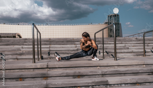 A girl in sportswear does a warm-up before jogging. Against the background of the stairs, in the summer in the city. Sports and healthy lifestyle. Tattoos on the body.