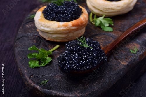 Black caviar in a wooden spoon and puff pastry tartlets on a dark board