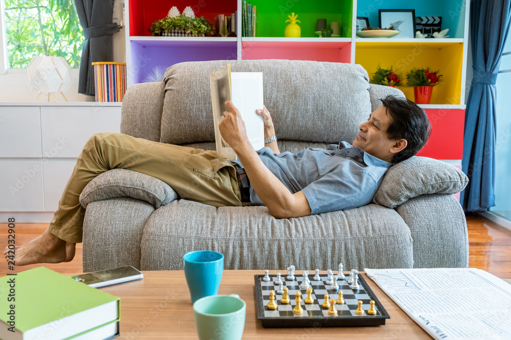 Obraz premium Asian middle-aged man lying on the sofa reading a magazine, very comfortable
