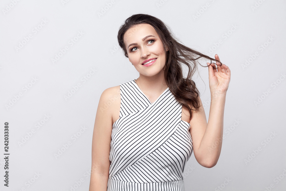 Happy optimistic beautiful young brunette woman with makeup and striped dress standing touching hair, dreaming and looking away with toothy smile. indoor studio shot, isolated on grey background. Happy optimistic beautiful young brunette woman with makeup and striped dress standing touching hair, dreaming and looking away with toothy smile. indoor studio shot, isolated on grey background.