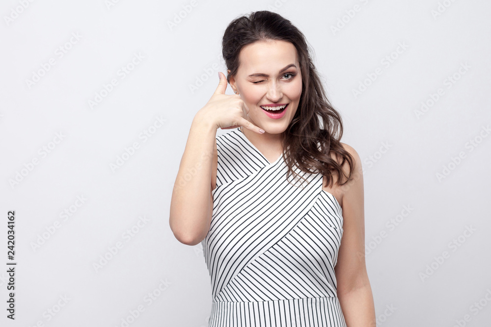 Call me. Portrait of funny beautiful young brunette woman with makeup and striped dress standing, winking and looking at camera with call gesture sign. indoor studio shot, isolated on grey background. Call me. Portrait of funny beautiful young brunette woman with makeup and striped dress standing, winking and looking at camera with call gesture sign. indoor studio shot, isolated on grey background.