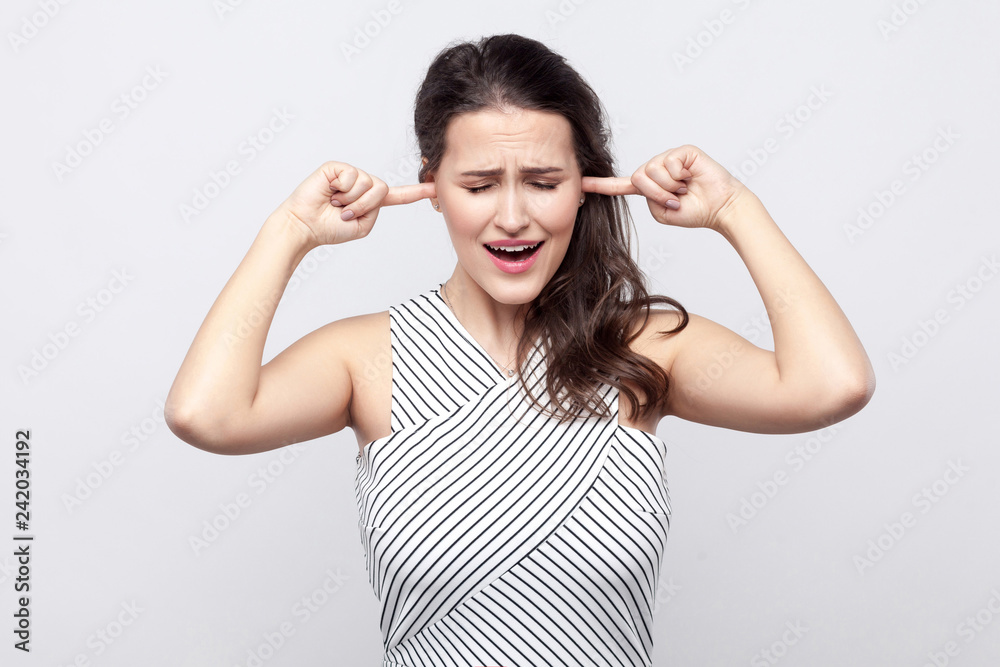 I don't want to here this. Portrait of unhappy beautiful young brunette woman with makeup and striped dress standing and holding fingers on her ear. indoor studio shot, isolated on grey background. I don't want to here this. Portrait of unhappy beautiful young brunette woman with makeup and striped dress standing and holding fingers on her ear. indoor studio shot, isolated on grey background.