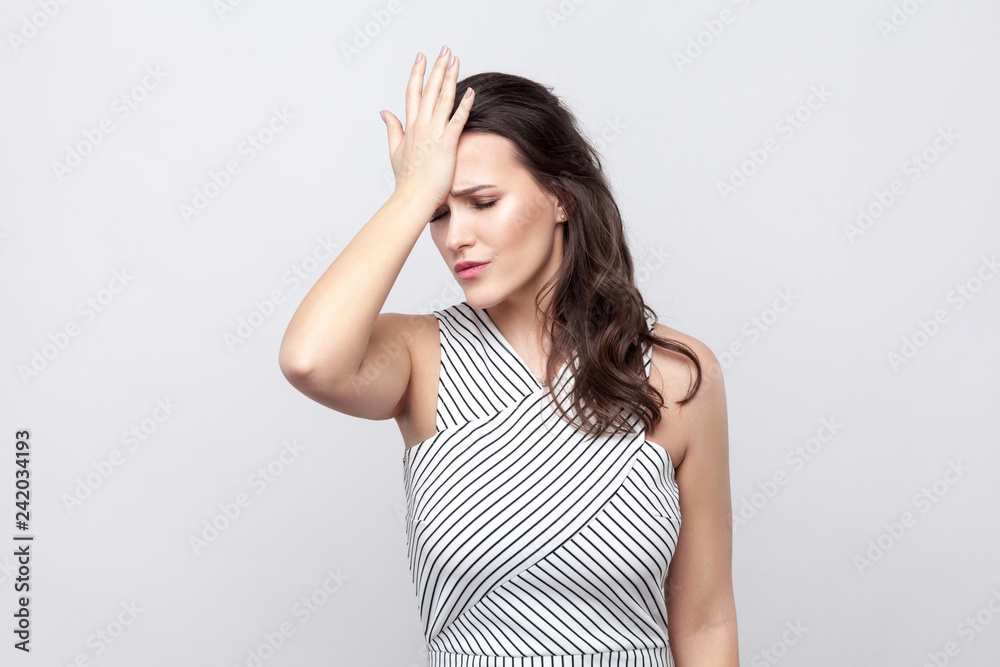 Portrait of sad lose beautiful young brunette woman with makeup and striped dress standing and holding hand on her forehead. indoor studio shot, isolated on grey background.