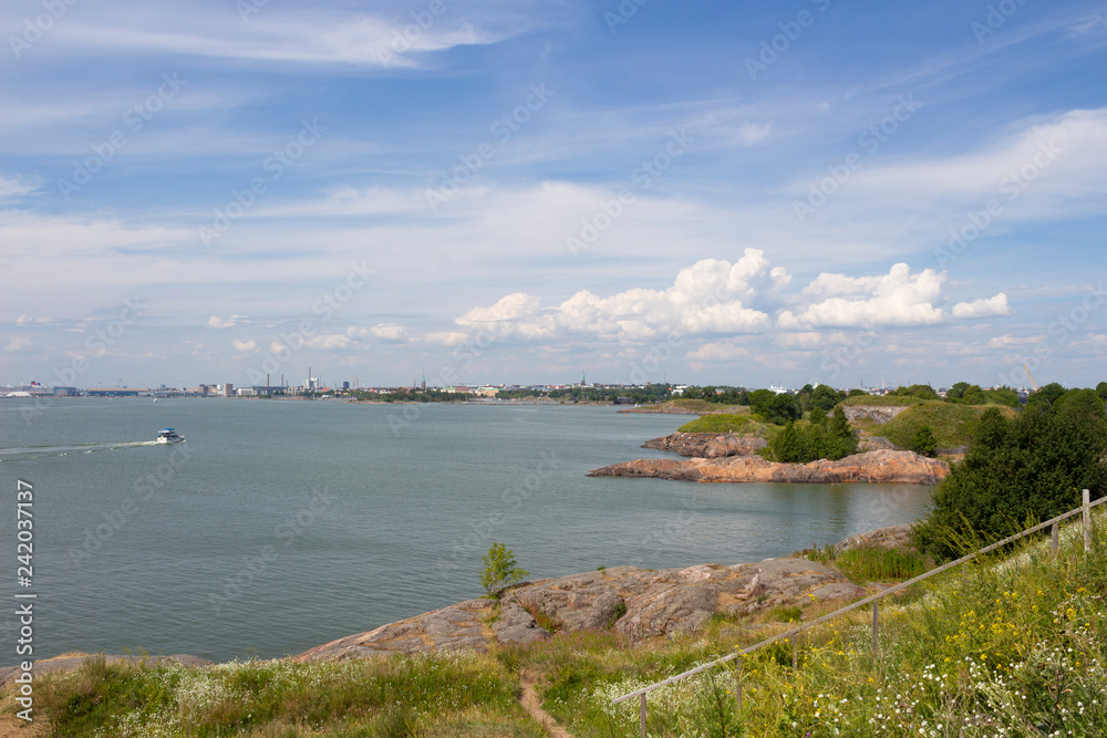 The stony shore of the island of Sumenlinna Sveaborg with grass and paths on it and a view of the Gulf of Finland and the city of Helsinki in the distance.