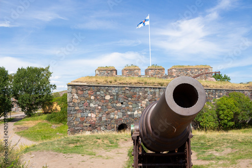 An old cast-iron artillery cannon and granite old fortifications on Suomenlinna fort island are overgrown with grass on a sunny summer day in Finland, and the flag of Finland is on the ancient walls.